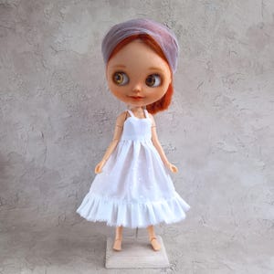 May include: A Blythe doll with red hair and a sheer, lavender head covering. The doll wears a white dress with a ruffled hem. The doll is standing on a small wooden stand against a textured gray background.