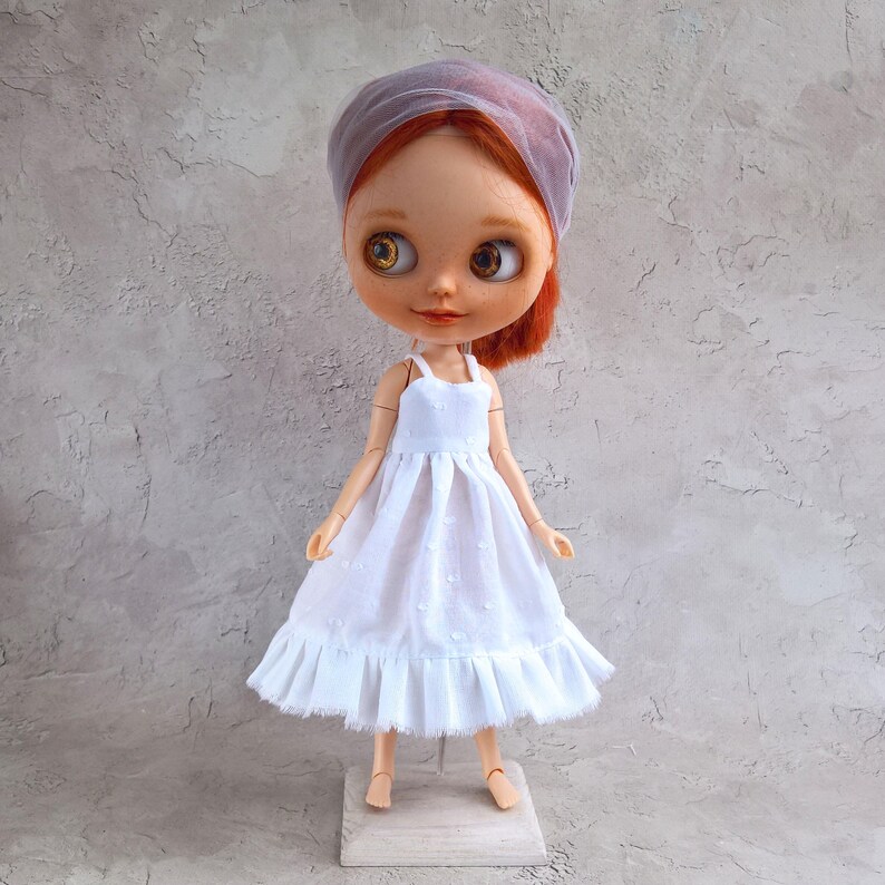 May include: A Blythe doll with red hair and a sheer, lavender head covering. The doll wears a white dress with a ruffled hem. The doll is standing on a small wooden stand against a textured gray background.