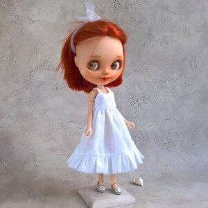 May include: A handmade doll with red hair and large, expressive eyes. The doll wears a white dress and silver shoes. A white ribbon adorns the doll's hair. The doll is standing on a small wooden block.