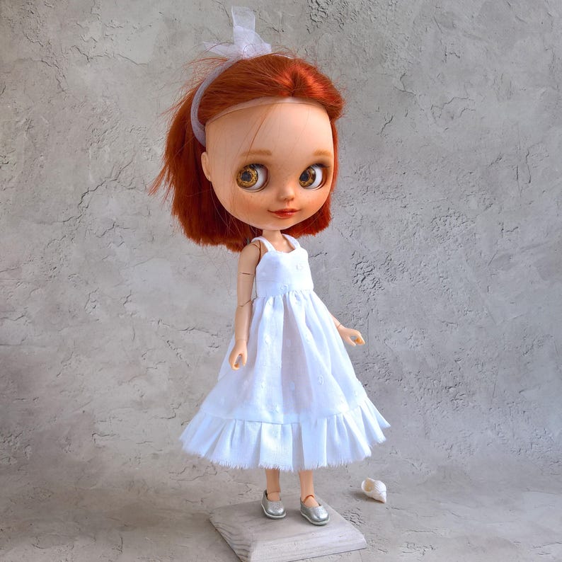 May include: A handmade doll with red hair and large, expressive eyes. The doll wears a white dress and silver shoes. A white ribbon adorns the doll's hair. The doll is standing on a small wooden block.