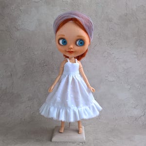 May include: A doll with red hair, blue eyes, and a white dress. The doll is wearing a sheer, light purple head covering. The dress has a ruffled hem and a polka dot pattern. The doll is standing on a small wooden stand.