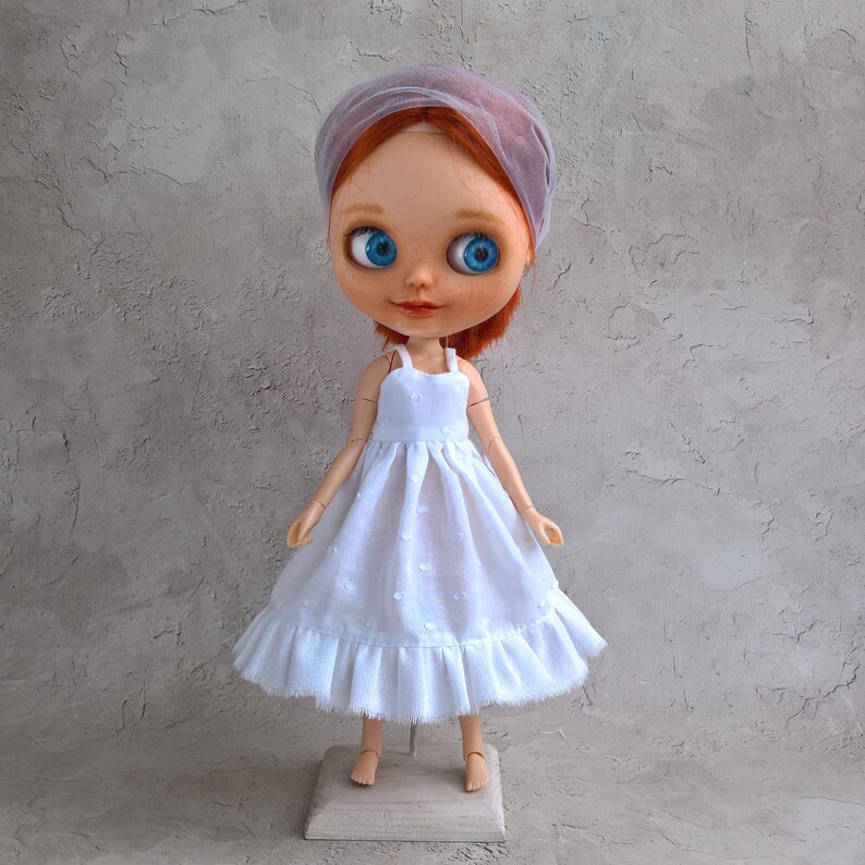 May include: A doll with red hair, blue eyes, and a white dress. The doll is wearing a sheer, light purple head covering. The dress has a ruffled hem and a polka dot pattern. The doll is standing on a small wooden stand.