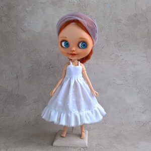 May include: A doll with red hair, blue eyes, and a light purple veil. The doll wears a white dress with a ruffled hem and a square base. The dress has a polka dot pattern.