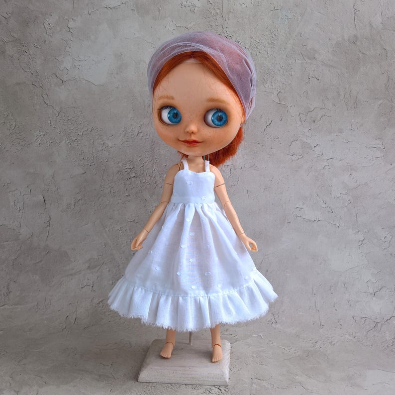 May include: A doll with red hair, blue eyes, and a light purple veil. The doll wears a white dress with a ruffled hem and a square base. The dress has a polka dot pattern.