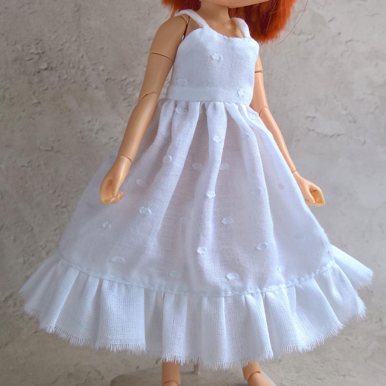 May include: A white doll dress with a textured, dotted pattern. The dress features thin straps, a fitted bodice, and a flared skirt with a ruffled hem. The doll dress is made of a lightweight fabric.