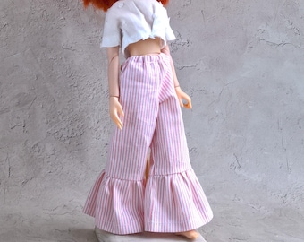 May include: A doll dressed in a white crop top and pink and white striped palazzo trousers with a ruffled hem. The doll has red hair and sunglasses. The doll is on a small, white, rectangular base.