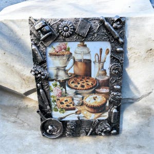 May include: A rectangular picture frame with a metallic silver finish, featuring a textured border decorated with miniature kitchen utensils and baking tools. The frame holds an image of various baked goods, including pies and pastries.