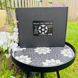 May include: A black rectangular box labelled Mosaic Side Table Kit with a list of contents including tiles, glue, grout, and a design print. The box sits on a round black metal side table featuring a grey and white floral mosaic tile pattern.