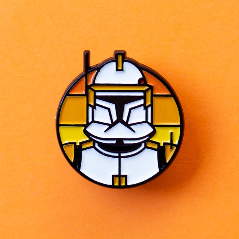 May include: A round enamel pin featuring a cartoon depiction of a Star Wars clone trooper with a white helmet and yellow and orange accents.