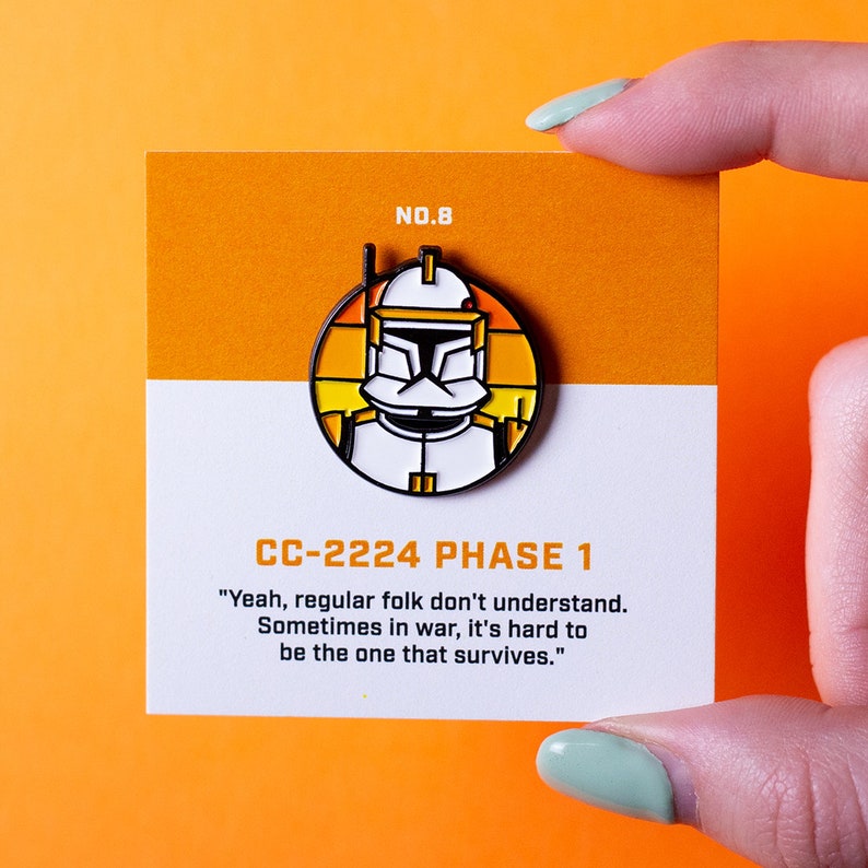 May include: A yellow and orange enamel pin featuring a Star Wars clone trooper with the text "CC-2224 PHASE 1" and the quote "Yeah, regular folk don't understand. Sometimes in war, it's hard to be the one that survives."