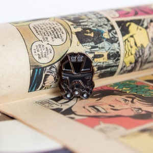 Op de afbeelding: Een zwarte en zilveren emaille pin in de vorm van een futuristische helm, rustend op een vintage stripboekpagina. De pin heeft een gedetailleerd ontwerp met drie cirkelvormige lichten en twee rechthoekige vormen aan de bovenkant. Het stripboek heeft kleurrijke panelen.