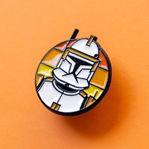 May include: A round enamel pin featuring a cartoon depiction of a Star Wars clone trooper with a white helmet and orange and yellow accents.