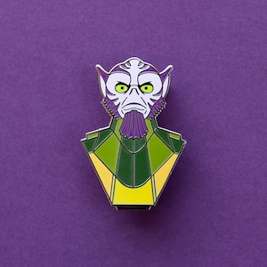 Puede incluir: Un pin de esmalte verde, amarillo y morado con un extraterrestre de dibujos animados con orejas grandes y una expresión seria.