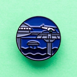 Op de afbeelding: Een blauwe en grijze emaille pin met een gestileerde illustratie van een gebouw met een klein bootje dat er voor aangemeerd ligt. Het gebouw heeft een plat dak en een reeks ramen. Het bootje is wit met een zwarte omtrek. De pin is rond en heeft een glanzende afwerking.