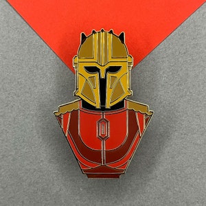 May include: A gold and red enamel pin featuring a stylised bust of a character wearing a helmet with horns. The helmet has a gold and black design with a red and gold chest plate.