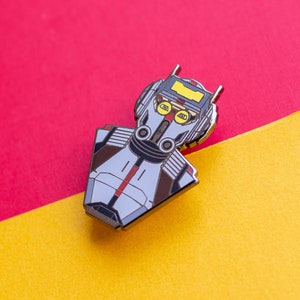 May include: A silver and black enamel pin featuring a robot character with a yellow visor and red eyes. The pin is on a red and yellow background.