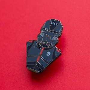 Pode incluir: Um broche de esmalte preto e prata que representa um personagem estilizado usando um capacete com o número "88" nele. O personagem está usando um terno escuro com um emblema de caveira no peito.