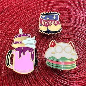 May include: Three enamel pins featuring a milkshake, a ghost wearing glasses, and a bag of Krisps crisps. The pins are gold-toned and have colourful details.