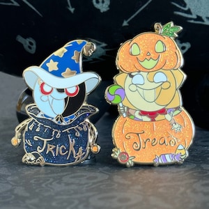 May include: Two enamel pins featuring Halloween characters. The first pin is a black and white wizard with a blue hat and a bag that says "Trick". The second pin is a pumpkin with a smiling face and a bag that says "Treat".