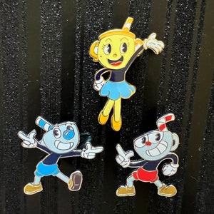 May include: Three enamel pins featuring characters from the video game Cuphead. The pins are yellow, blue, and red. The characters are Cuphead, Mugman, and Ms. Chalice.