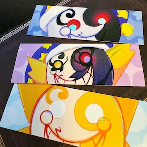 May include: Three colorful bookmarks featuring a cartoon character with a black and white face. The character has a large, toothy grin and a star-shaped hat. The bookmarks are printed on cardstock and have a glossy finish.