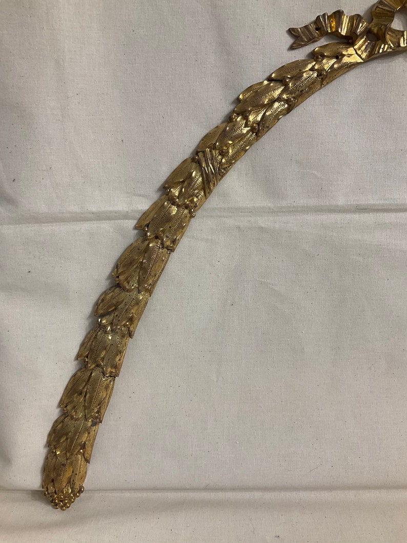 May include: A gold-colored metal decorative trim with a leaf design. The trim is curved and has a ribbon at the top.
