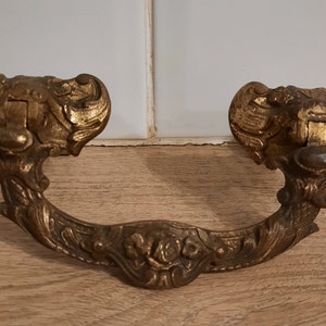 Pode incluir: Uma puxador de gaveta de metal dourado com um design ornamentado que apresenta motivos florais e um anel central. O puxador tem uma estética vintage e seria uma adição elegante a qualquer móvel.