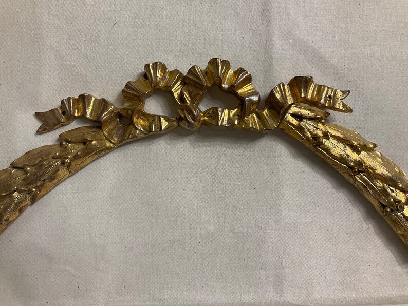 May include: A gold-colored metal frame with a decorative ribbon design. The frame is curved and has a floral pattern.
