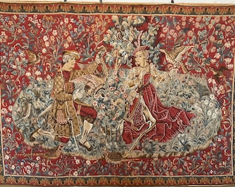 May include: A tapestry depicting a man and woman in a garden setting. The tapestry features a red background with intricate floral patterns and birds. The man is wearing a brown and green outfit, while the woman is wearing a red dress. The tapestry is framed in a gold and black border.