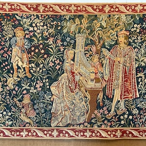 Puede incluir: Un tapiz que representa una escena medieval con figuras en un entorno de jardín. El tapiz presenta a una mujer tocando un instrumento musical, un hombre cantando y una pareja de pie cerca de un árbol. El tapiz está tejido con intrincados detalles y colores vibrantes, incluyendo tonos de verde, azul, rojo y dorado.