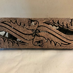 May include: A wooden batik printing block with a carved design of stylized floral patterns. The block is brown and has a rectangular shape.