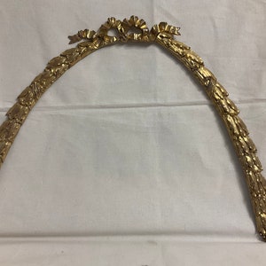 May include: A gold-colored metal decorative piece shaped like an archway with a leafy design and a bow at the top.