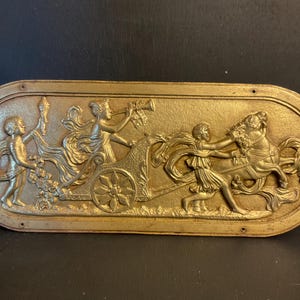 Könnte beinhalten: Eine ovale, goldfarbene Plakette mit einer erhabenen Reliefskulptur, die einen von einem Pferd gezogenen Wagen darstellt. Der Wagen wird von einer Frau gefahren, die eine Trompete hält, und eine zweite Frau hält die Zügel. Eine dritte Figur steht hinter dem Wagen und hält eine Fackel.