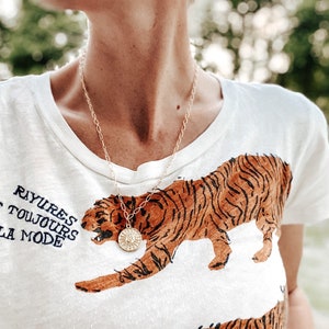 May include: A gold-colored necklace with a circular pendant and a white t-shirt featuring a tiger graphic in shades of brown and black. The shirt also has French text that reads "Rayures sont toujours à la mode."