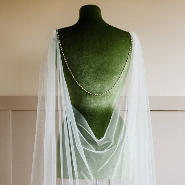 May include: A sheer white tulle capelet draped over a green velvet mannequin torso. The capelet features a delicate gold chain adorned with small white beads that connects across the back, with long, flowing tulle panels extending downward.