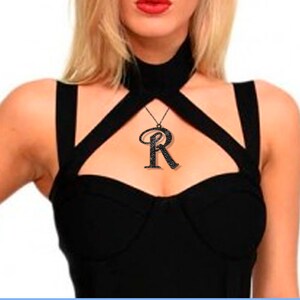 K&ouml;nnte beinhalten: Eine schwarze Choker-Kette mit einem gro&szlig;en, schwarzen, mit Strasssteinen besetzten Anh&auml;nger in Form des Buchstabens "R".