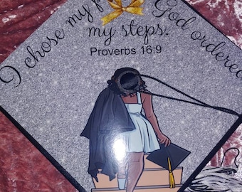 May include: A graduation cap with a silver glitter background and a black silhouette of a person wearing a graduation gown and cap walking up a staircase. The cap has a gold bow and the text "I chose my path but God ordered my steps. Proverbs 16:9."