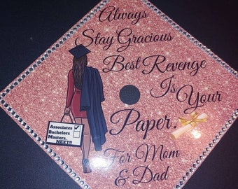 May include: A pink and gold graduation cap with rhinestones around the edge. The cap features a black and white illustration of a woman in a graduation gown and cap, holding a bag and a diploma. The text on the cap reads "Always Stay Gracious Best Revenge Is Your Paper. For Mom & Dad."