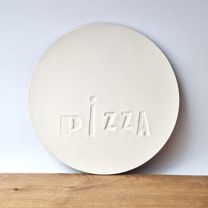 Könnte beinhalten: Ein runder, cremefarbener Pizzastein mit dem geprägten Wort "PIZZA". Der Stein liegt auf einem Holzschneidebrett mit natürlicher Holzmaserung. Der Hintergrund ist schlicht weiß.