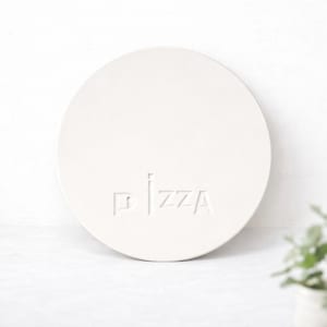 Könnte beinhalten: Ein runder, weißer Pizzastein mit dem Wort "PIZZA" auf der Oberfläche geprägt. Der Stein hat eine einfarbige, neutrale Farbe, und der Text ist dezent in das Design integriert. Der Stein ist zum Backen von Pizzen bestimmt.