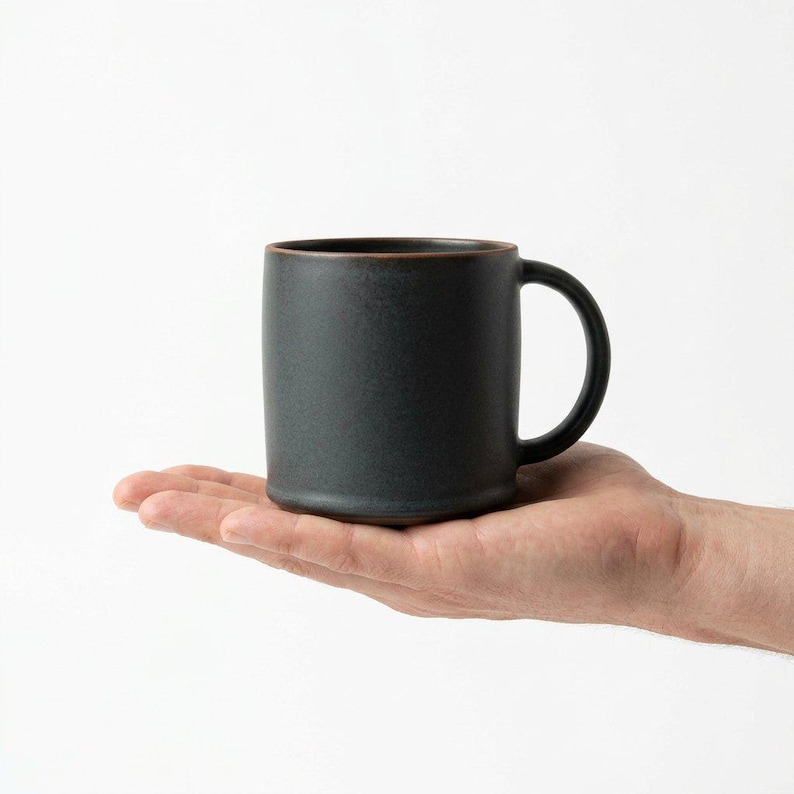 May include: A dark gray ceramic mug with a rounded handle and a brown rim. The mug is held in a person's hand against a white background. The mug is approximately 4 inches tall.
