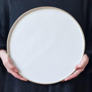 Könnte beinhalten: Ein großer, weißer Keramik-Servierteller mit leicht erhöhtem Rand. Der Teller ist rund und hat eine glatte, matte Oberfläche.
