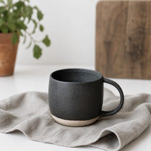 Könnte beinhalten: Eine dunkelgraue Keramiktasse mit rundem Körper und Henkel. Der Boden der Tasse ist hellbeige. Die Tasse steht auf einer grauen Leinen-Serviette. Eine Topfpflanze und ein Holzschneidebrett befinden sich im Hintergrund.