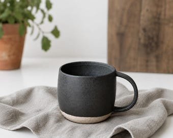 May include: A dark gray ceramic mug with a rounded body and a handle. The bottom of the mug is a light beige color. The mug sits on a gray linen napkin. A potted plant and a wooden cutting board are in the background.