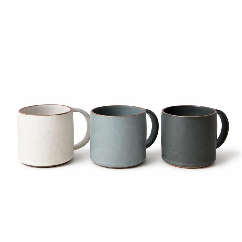 May include: Three ceramic mugs in a row, each with a rounded handle. The mugs are in shades of white, blue-grey, and dark grey. The rims and bases have a contrasting brown color.