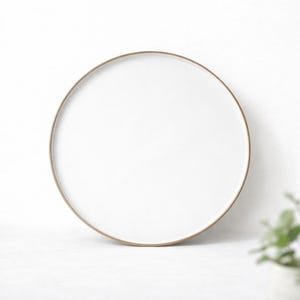 Könnte beinhalten: Ein runder, weißer Keramikteller mit einem dünnen, hellbraunen Rand. Der Teller ist vor einem weißen Hintergrund zentriert. Der Teller hat ein schlichtes, minimalistisches Design und eignet sich zum Servieren von Speisen.