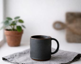 May include: A dark gray ceramic mug with a brown rim and base, filled with a dark liquid. The mug sits on a gray linen napkin. A small potted plant and a wooden cutting board are in the background.