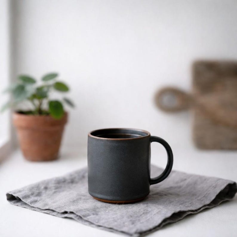 May include: A dark gray ceramic mug with a brown rim and base, filled with a dark liquid. The mug sits on a gray linen napkin. A small potted plant and a wooden cutting board are in the background.