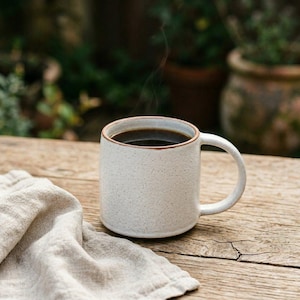 May include: A white speckled ceramic mug filled with dark coffee, with steam rising from the top. The mug has a rounded handle and a brown rim. A beige linen napkin rests on a wooden surface.