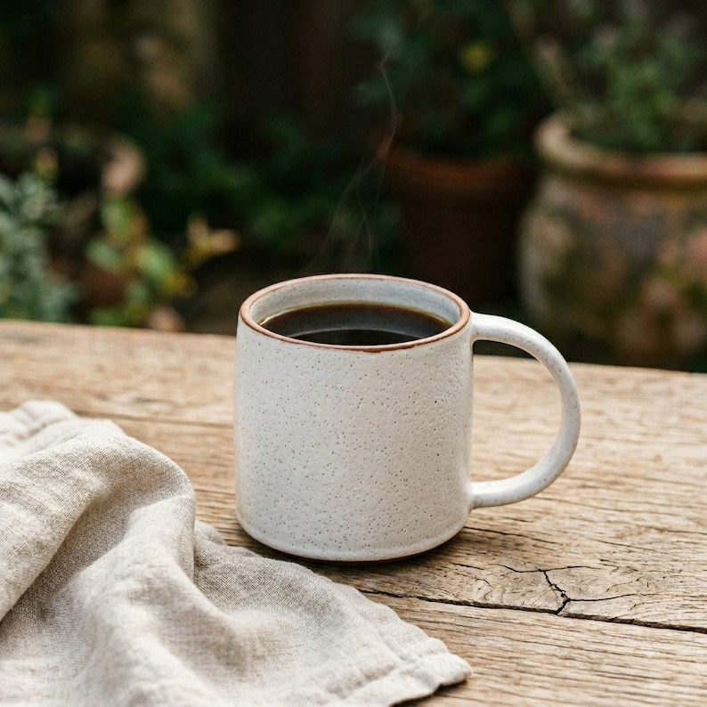 May include: A white speckled ceramic mug filled with dark coffee, with steam rising from the top. The mug has a rounded handle and a brown rim. A beige linen napkin rests on a wooden surface.